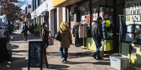 People walk down a city street outside a sign that says ASTORIA FOOD PANTRY