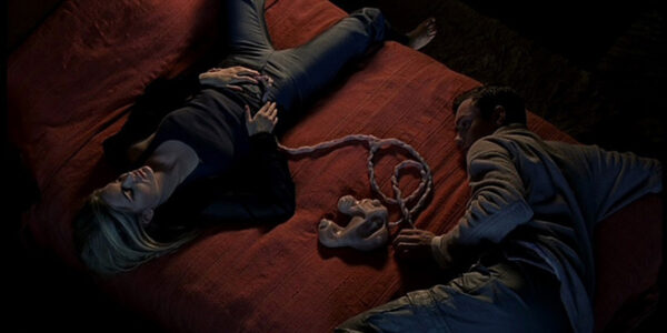 A man and woman lie on a bed apart from each other, connected by fleshy umbilical-like cords in their stomachs