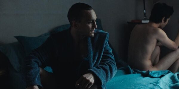 A man wearing a coat sits on a bed turning his head towards a naked man on the other side of the bed, turned away