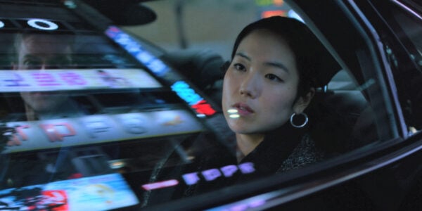 A woman looks out the window from the back seat of a car, with neon city lights reflected in the window