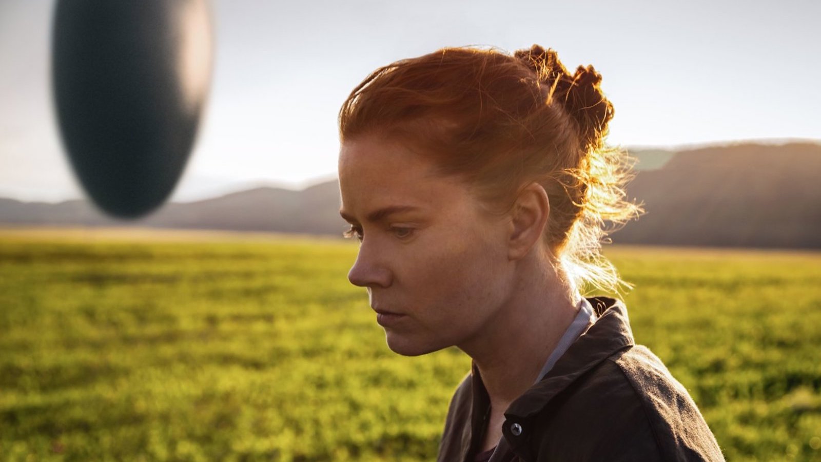 A woman stands in a field in profile with a hovering spacecraft behind her in the background