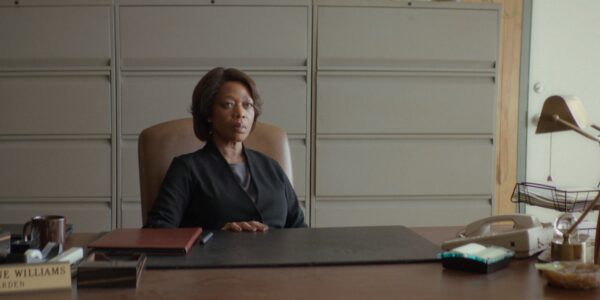 A woman with a serious expression and a black suit sits behind a desk in a drably colored office
