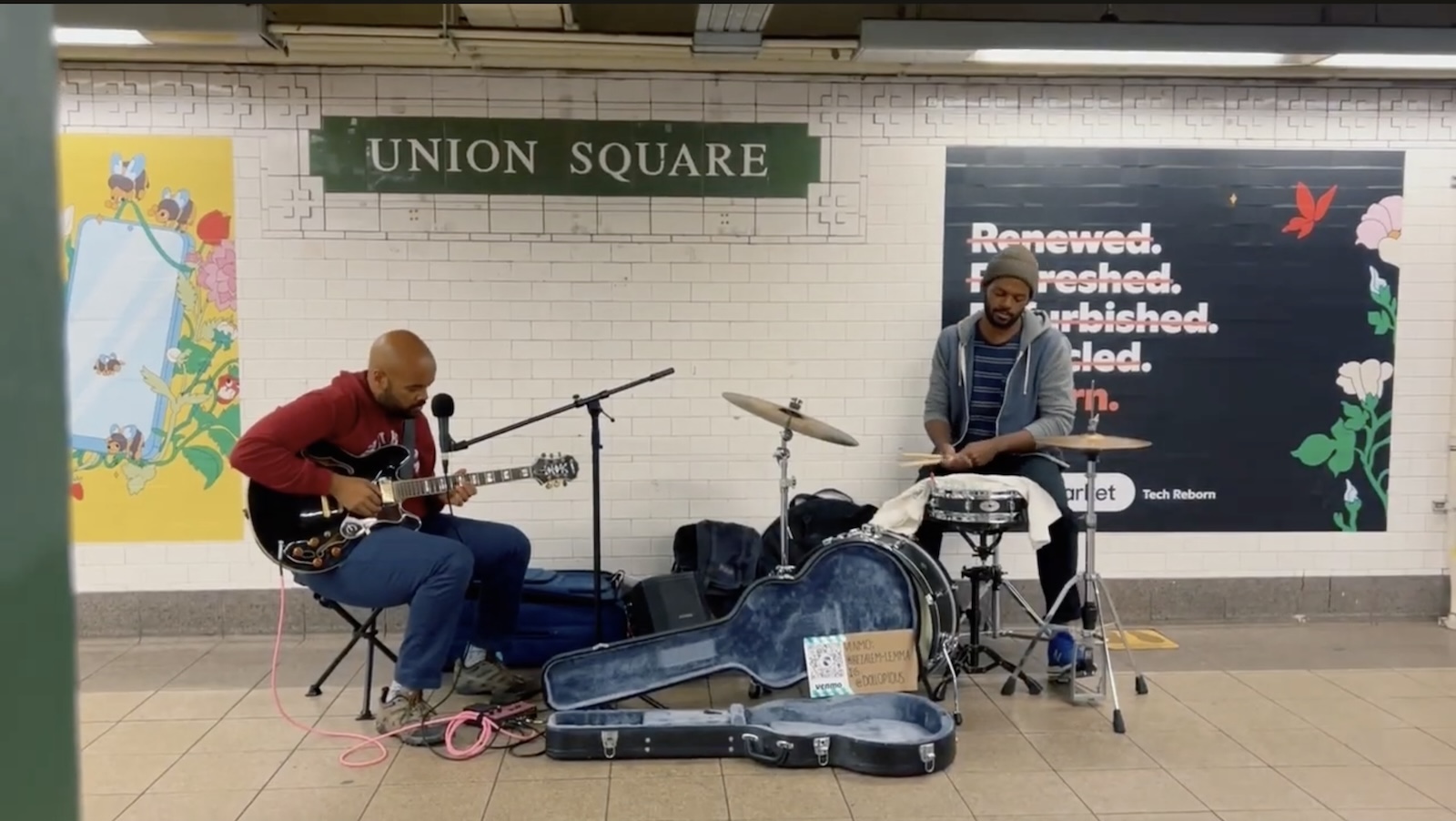 Two men play drums and guitar in the subway with the sign UNION SQUARE behind them on the wall
