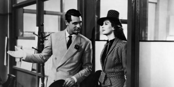 A man in a gray suit welcomes a woman in a pin-striped suit and hat into a newspaper office in a black and white photo