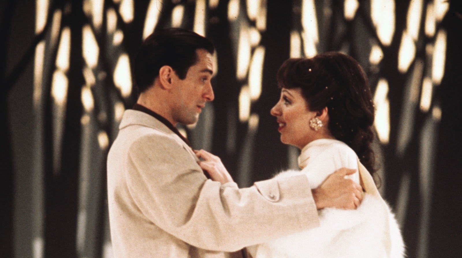 A man in a beige suit and a woman in a white fur coat stand in profile and look at each other, their hands gripping each other, against a forest backdrop