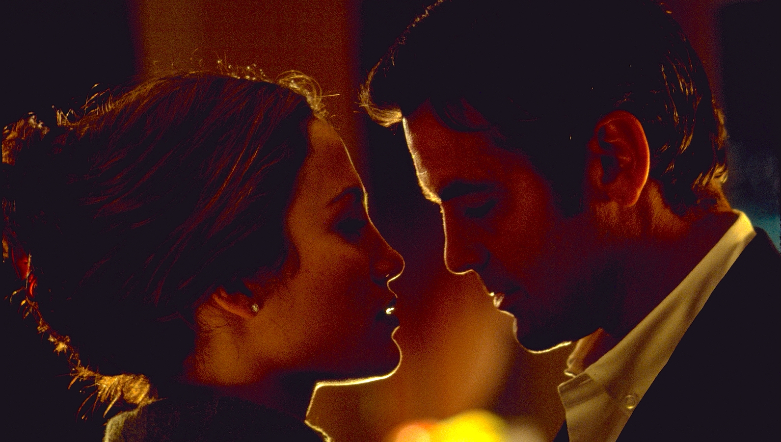 A close up of a man and woman looking at each other in a dark room in profile, ready to kiss