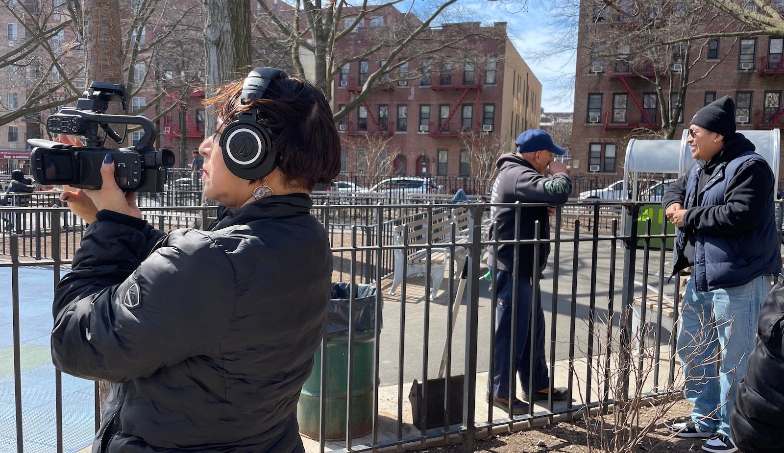 A woman holds a camera and wears headphones outside in New York, filming over an iron fence.
