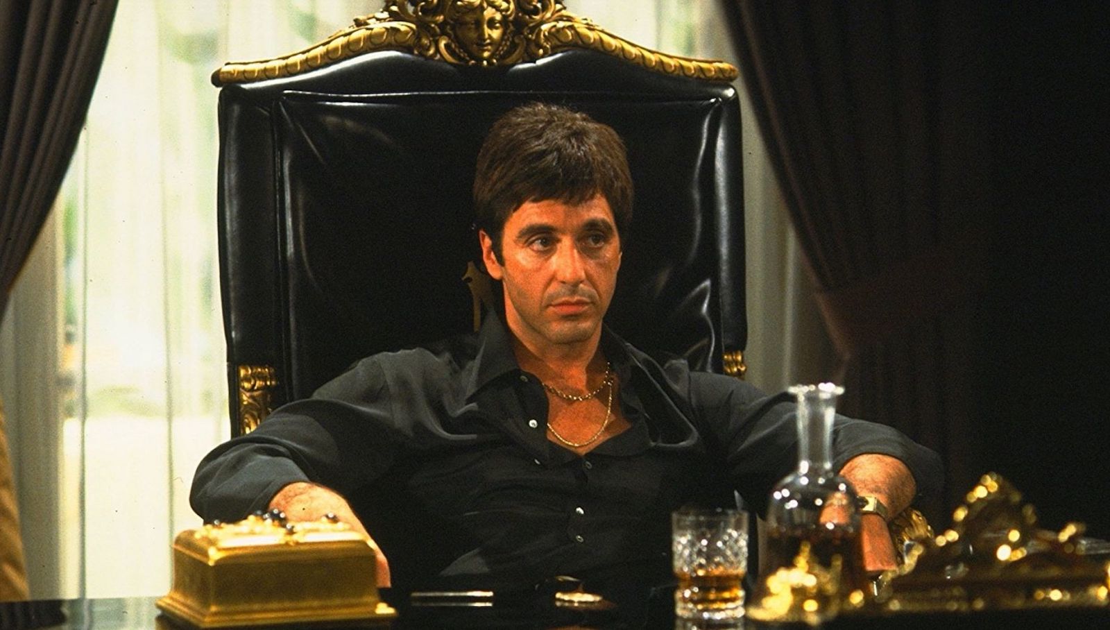 A man with a silver necklace and open collar sits in an ornate gold and black chair behind a desk in front of a whisky decanter and glass