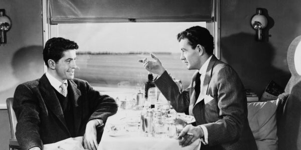 Two men sit at a table on a train compartment, talking pleasantly while landscape goes by in the back of the black and white image