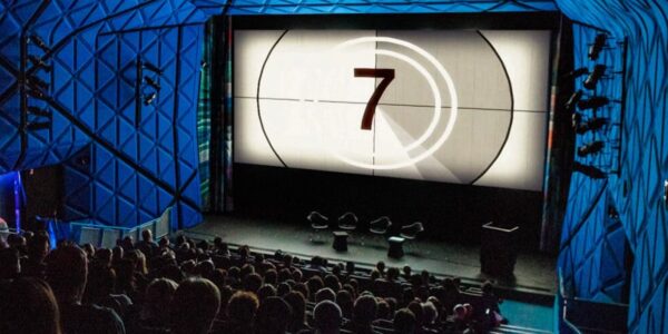A movie theater with a full audience looking at a screen with the number 7 on it counting down to showtime