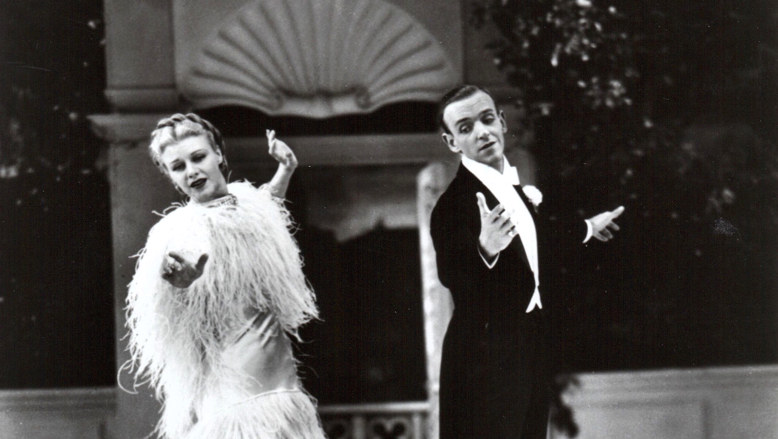 In a black and white photo a man in a tuxedo and a woman in a fluttering feathery dress dance side by side facing the camera, each with an arm outstretched