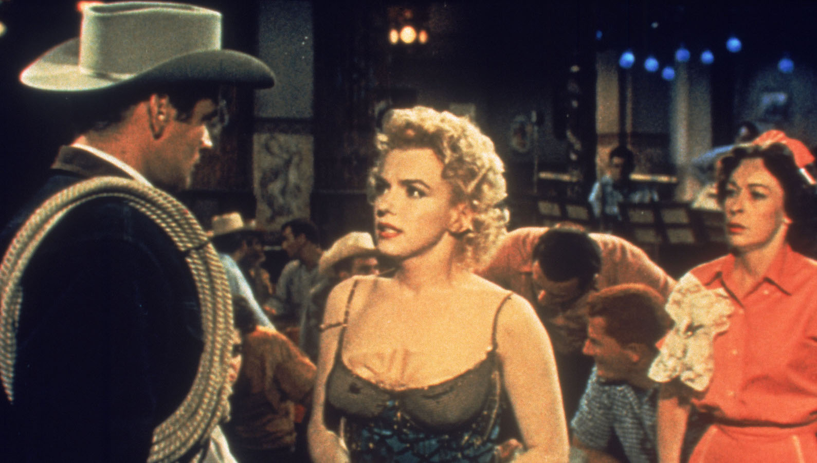 A blonde woman in a low-cut brown top talks intensely to a man in a cowboy hat and holding a coiled lasso rope over his shoulder