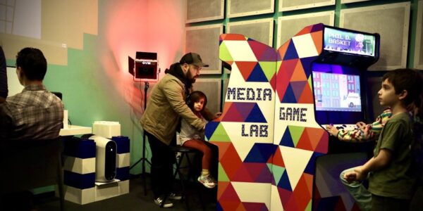 A father and daughter sitting at a video game arcade console; the side of the arcade console says Media Game Lab.