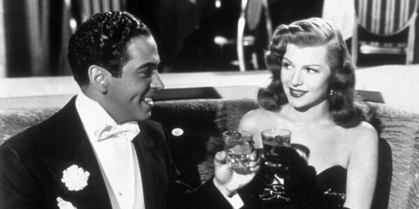 A man in a tux and a woman in a black, sleeveless dress toast glasses in a fancy restaurant in a black and white image