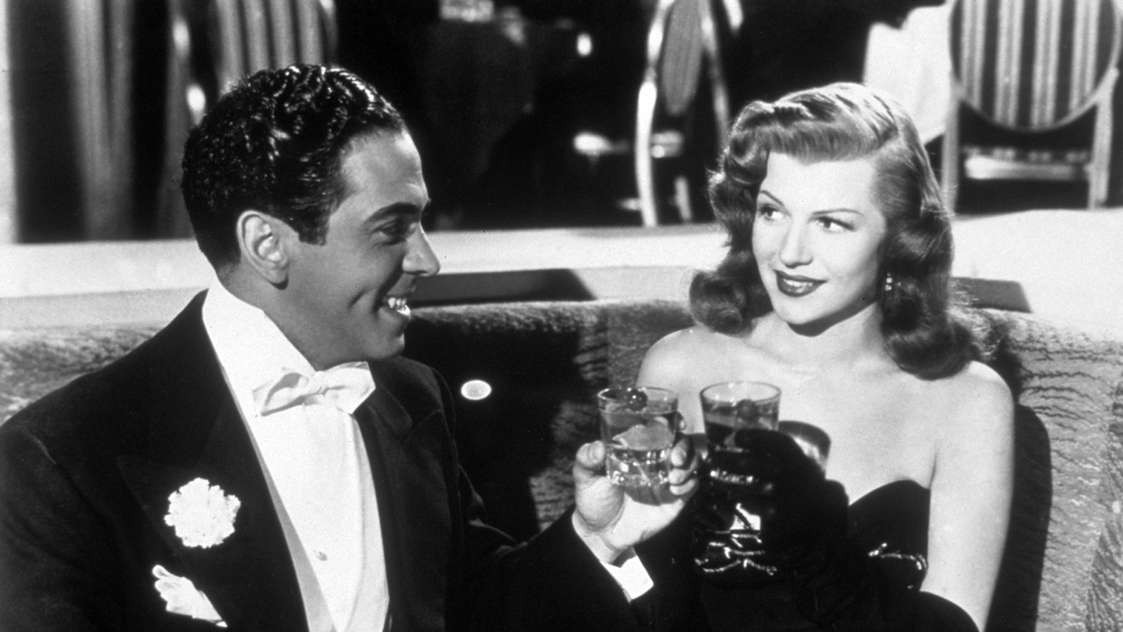 A man in a tux and a woman in a black, sleeveless dress toast glasses in a fancy restaurant in a black and white image