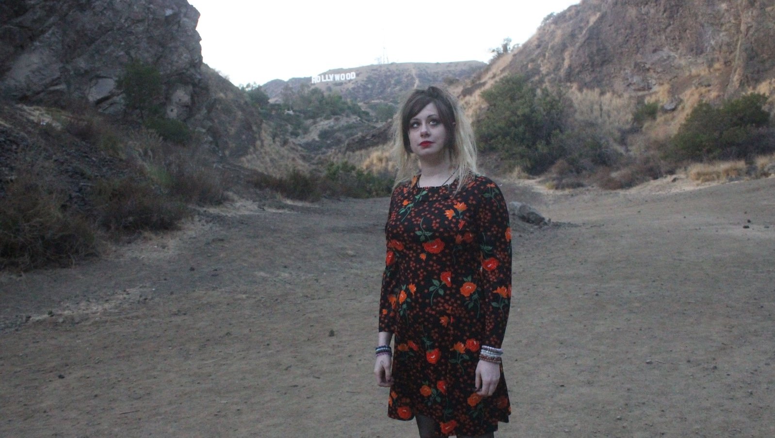 A girl in a red flowery dress and dyed blonde hair looks up while standing at the beach, which the HOLLYWOOD sign far in the background distance