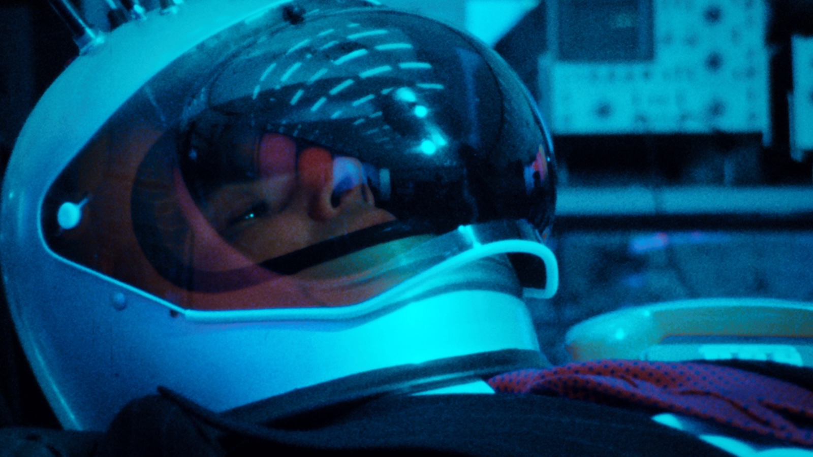 A man in a space helmet reclines, with reflections of lights on the glass viser