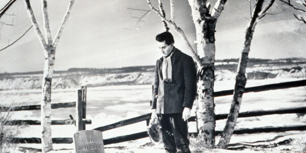 Against a snowy landscape, a man stands in a cemetery looking down sadly at a gravestone in black and white image