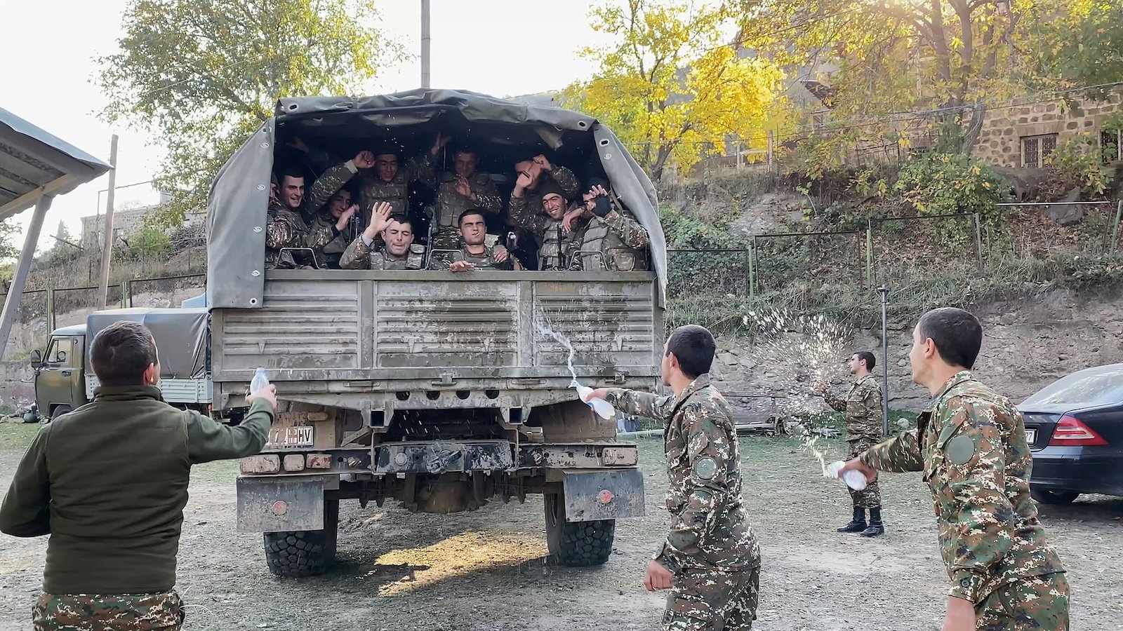 A truck full of soldiers drives away, while men dressed in fatigues wave from the back of the truck as four other men say goodbye to them by splashing bottled water at them.