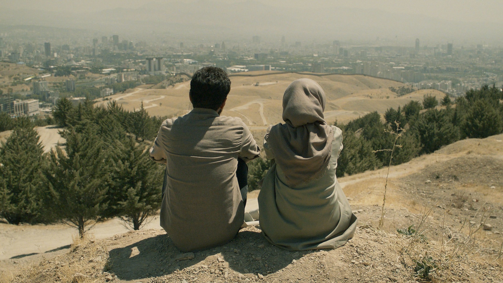 Seen from behind, a man and woman sit on a hillside looking over a parched, dusty middle-eastern landscape