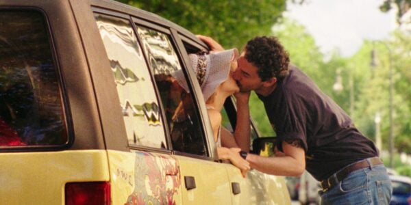 A woman sticks her head out the passenger side of a parked car, kissing a man standing next to the car in a passionate embrace