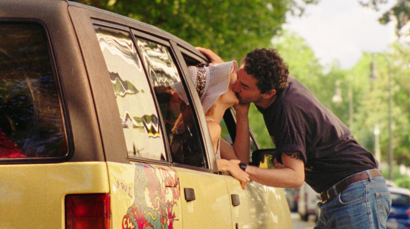 A woman sticks her head out the passenger side of a parked car, kissing a man standing next to the car in a passionate embrace