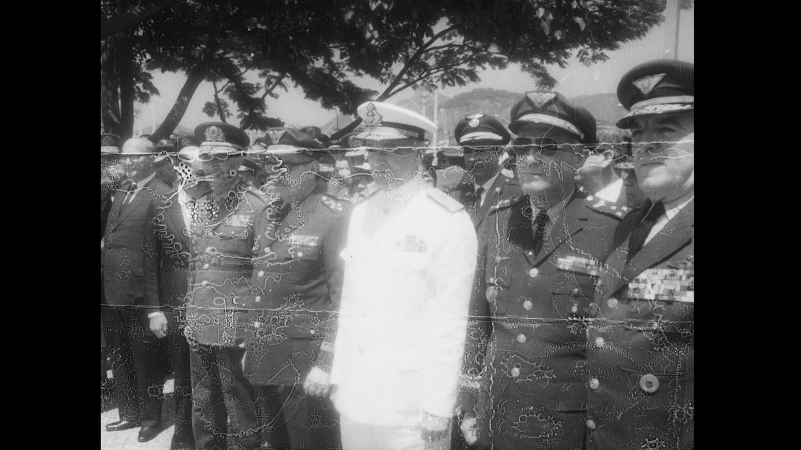 A black and white archival photo of military generals in uniform, one wearing white, the others darker uniforms, all wearing hats; the image is spotted with water damage and wear and tear.