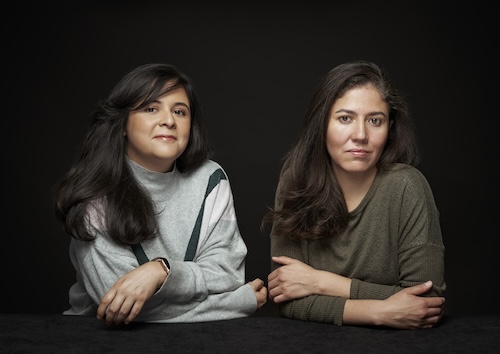 Two women cross their arms side by side and look at camera amidst a black background