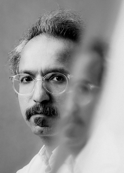 A portrait image of a bespectacled man, his face refracted in a mirror next to him