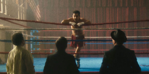 A boxer leans on the ropes in a boxing ring and talks to three men standing on the floor below looking up at him.