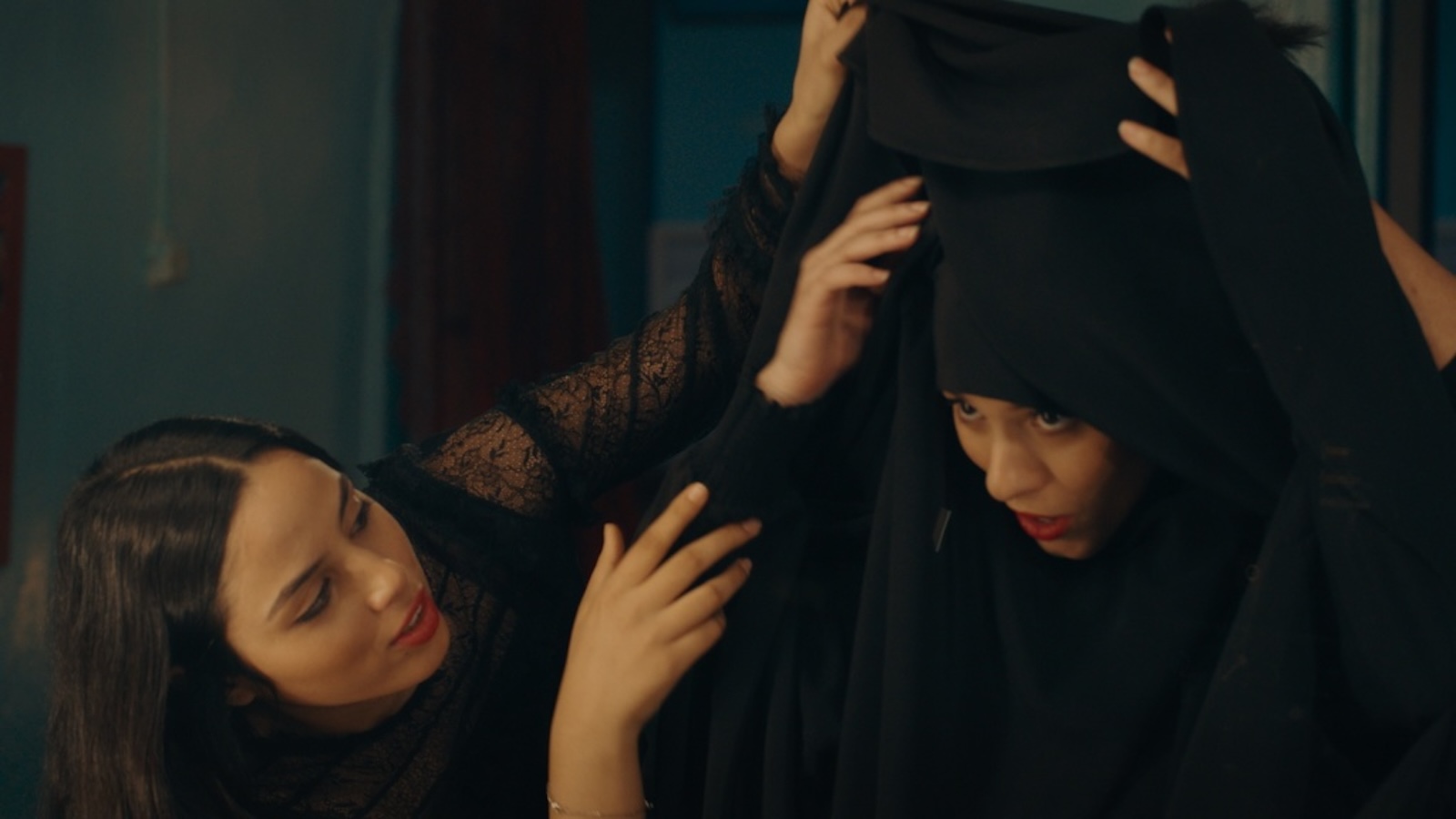 A woman helps another woman put on a black hijab.
