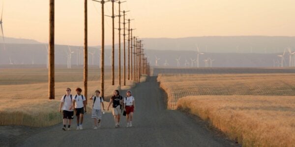 A group of teenagers walk down a vast American heartland landscape dotted with windmills