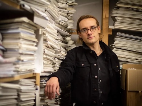 A man poses for the camera next to shelves and stacks of papers