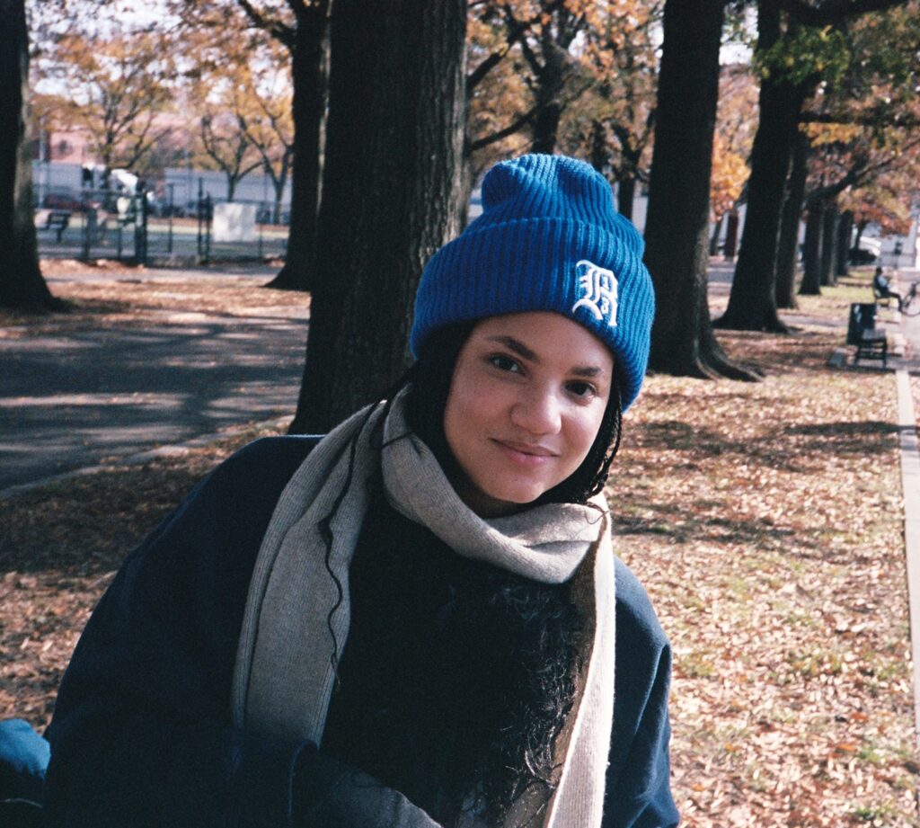 A person wearing a winter hat and scarf smiles in front of trees