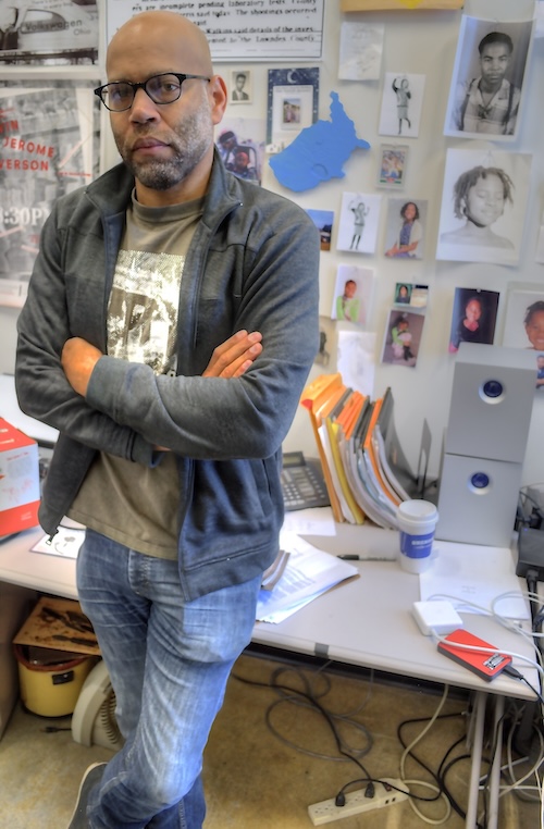 A man crosses his arms and looks at camera standing near a messy desk and wall with pictures taped to it