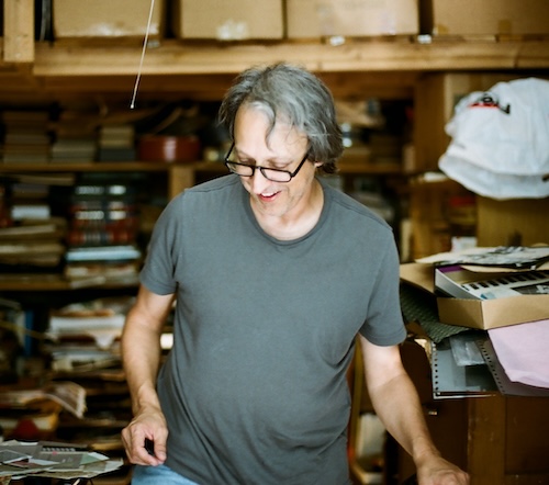 A person in a gray t-shirt stands in a cluttered room, smiling as he looks down