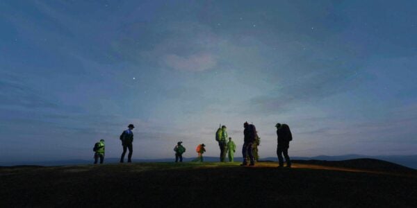 Eight human-sized figures seen from a distance standing against a vast blue sky horizon at twilight with some stars just coming out
