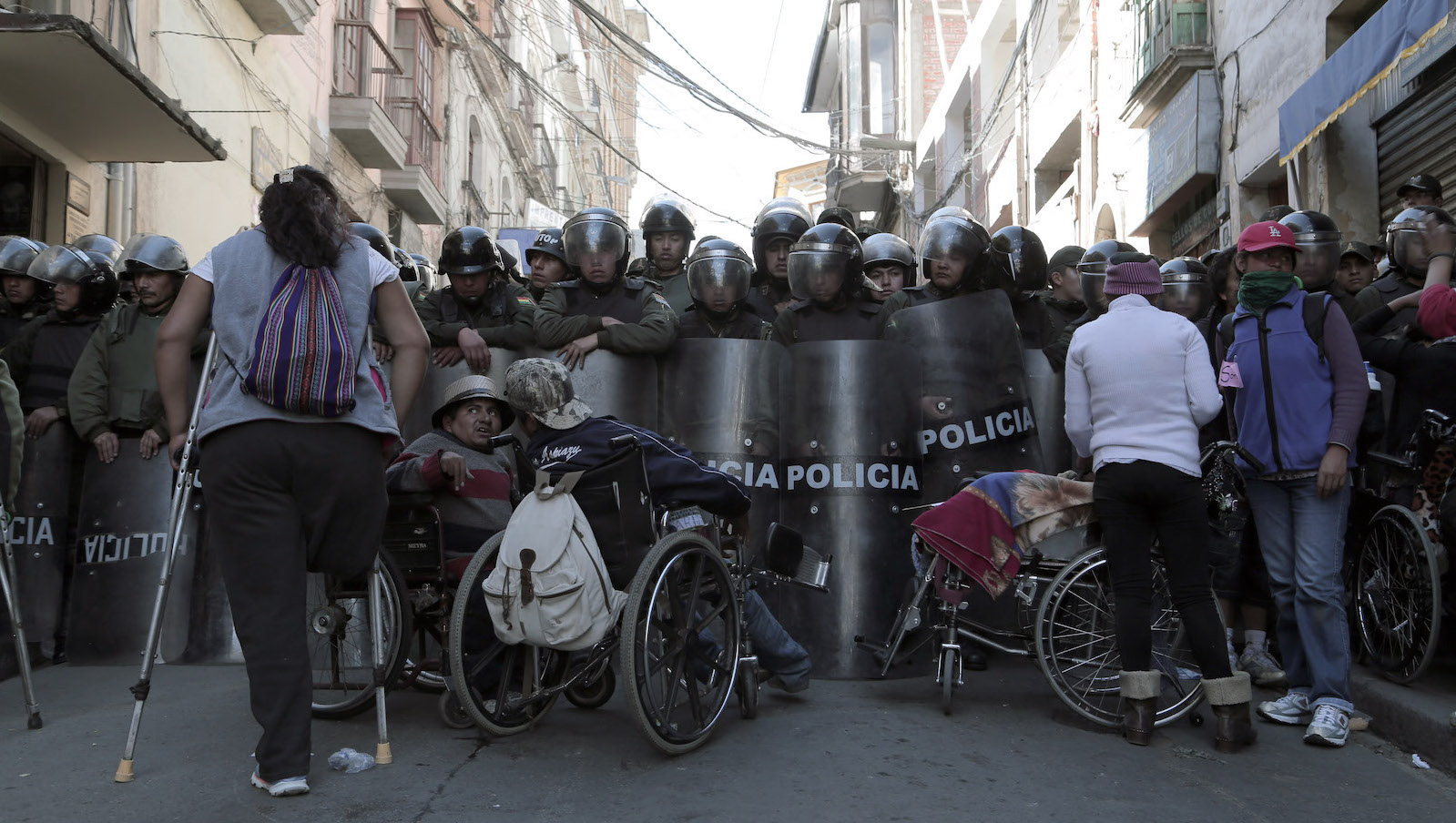 A group of disabled protestors in wheelchairs and one woman with crutches stands in front of a phalanx of police in riot gear and shields