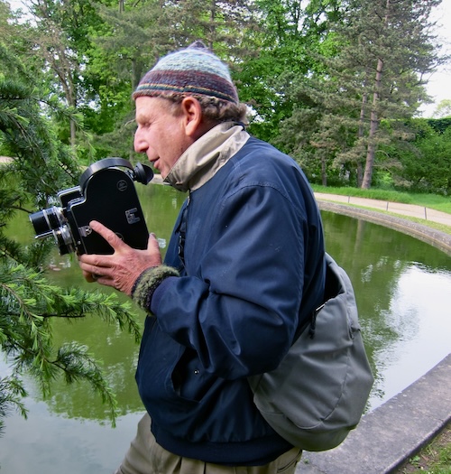 A man in a knit cap stands near a fountain pool of water holding a film camera