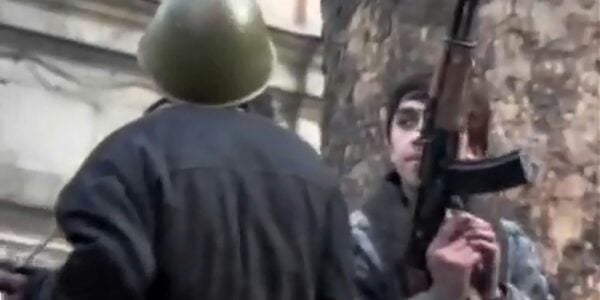 A degraded color video image of two men, one holding an semiautomatic rifle, covering half his face, the other from behind wearing an army helmet
