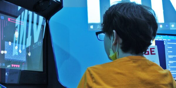 A young person wearing glasses, a yellow shirt, and a facemask dangling from their ears, sits in front of an arcade game, their back to the camera.