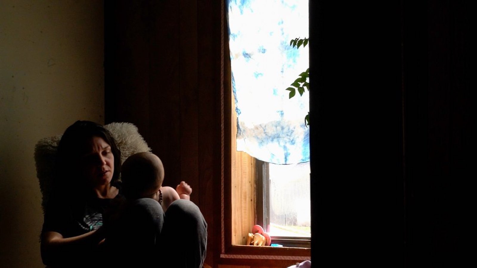 A woman holds a baby in a dark room near a window with daylight pouring in