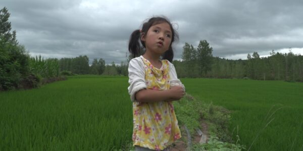 A little girl stands with her arms crossed against a vast green landscape