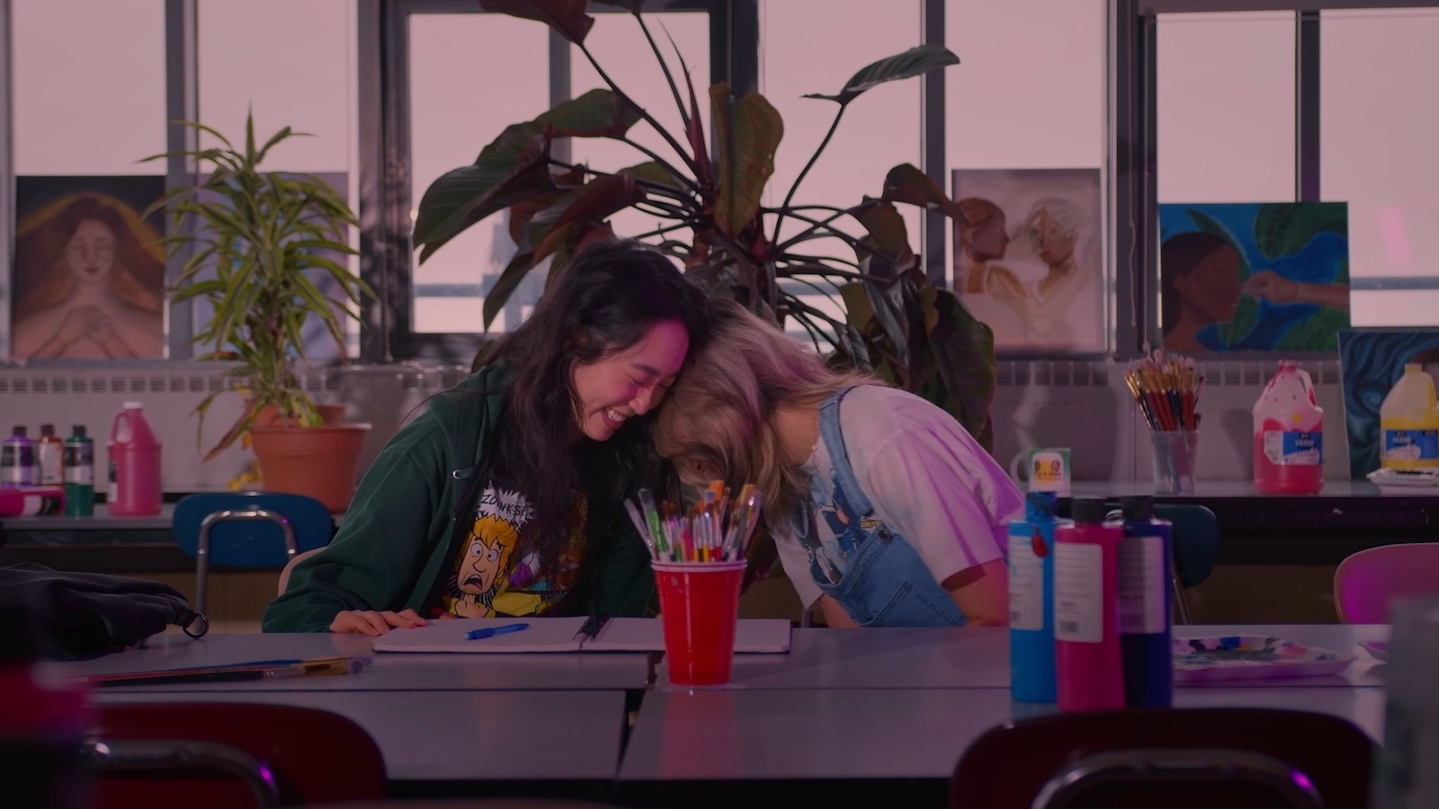 Two friends smile and laugh while sitting in a school room in front of a craft table, their heads on each other's shoulders