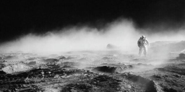 A man in a space suit seen from a distance walks across a rocky terrain enshrouded in fog