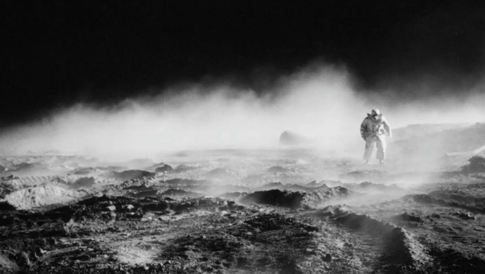 A man in a space suit seen from a distance walks across a rocky terrain enshrouded in fog