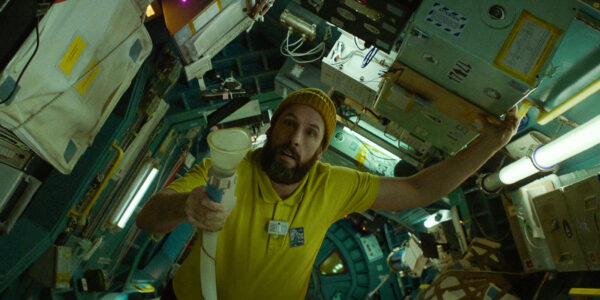A bearded man in a yellow shirt and knit cap stands in a spaceship with an alarmed look on his face, holding a vaccuum-like tube and nozzle as he looks up.