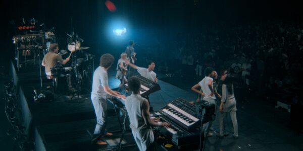 An image of a concert filmed from behind the musicians, featuring two keyboardists, drums, a soloist with one hand up and holding a.microphone, and a group of three backup singers.