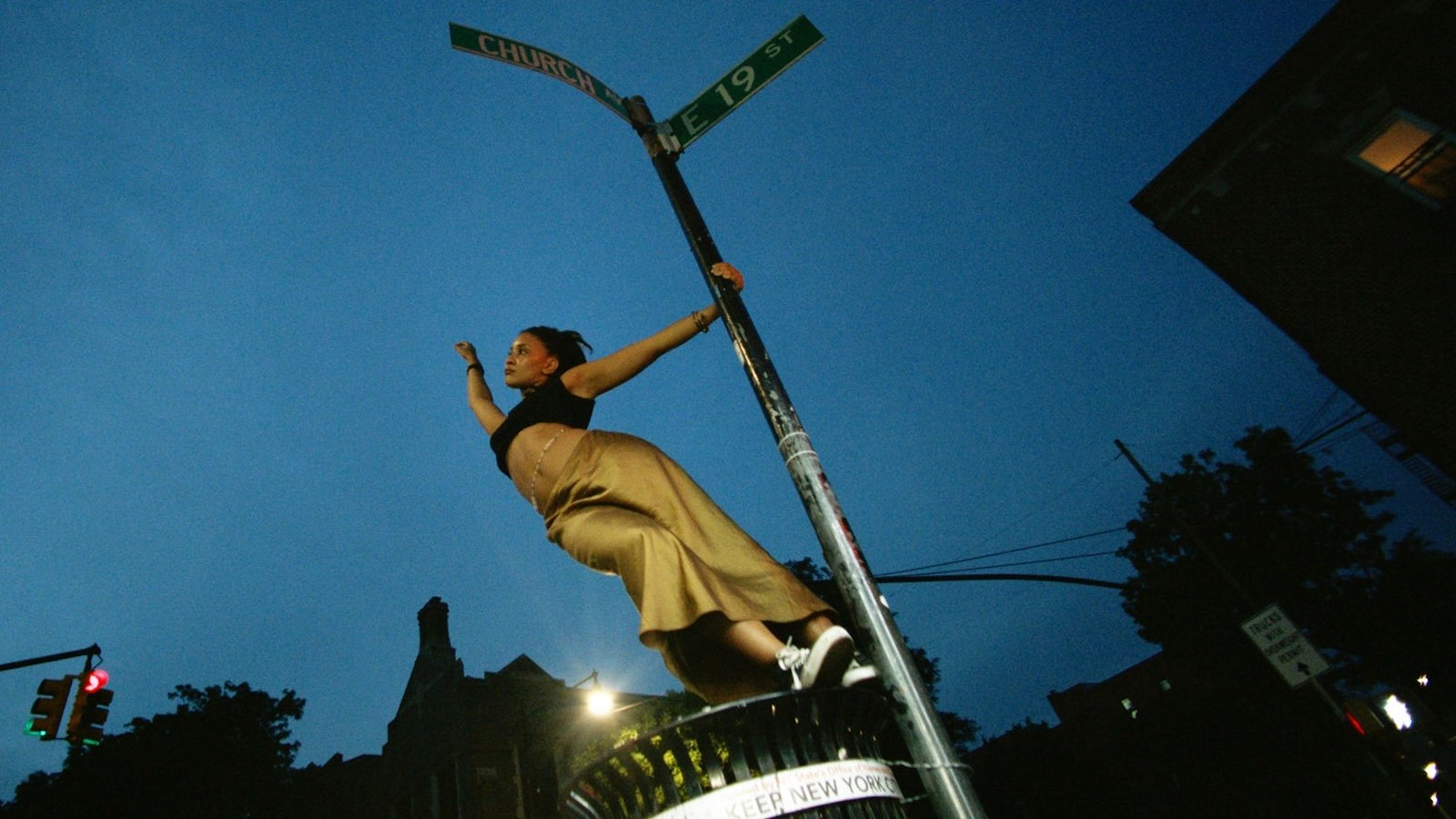A young woman bearing her midriff stands on a garbage can and grasping a New York city street sign, with one arm out
