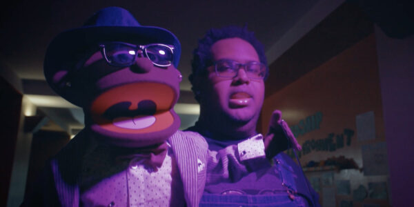 A person holds a puppet in the foreground, with a purple light shining on them; the puppet wears a hat and sunglasses and its mouth is open